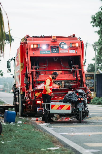 A worker wearing a high-visibility yellow vest with reflective stripes, black sleeves, and dark trousers is operating a waste collection vehicle on a city street during evening or dusk. The worker, wh